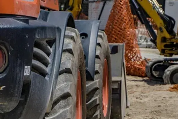 Construction vehicles on a dirt site with orange safety netting in background.
